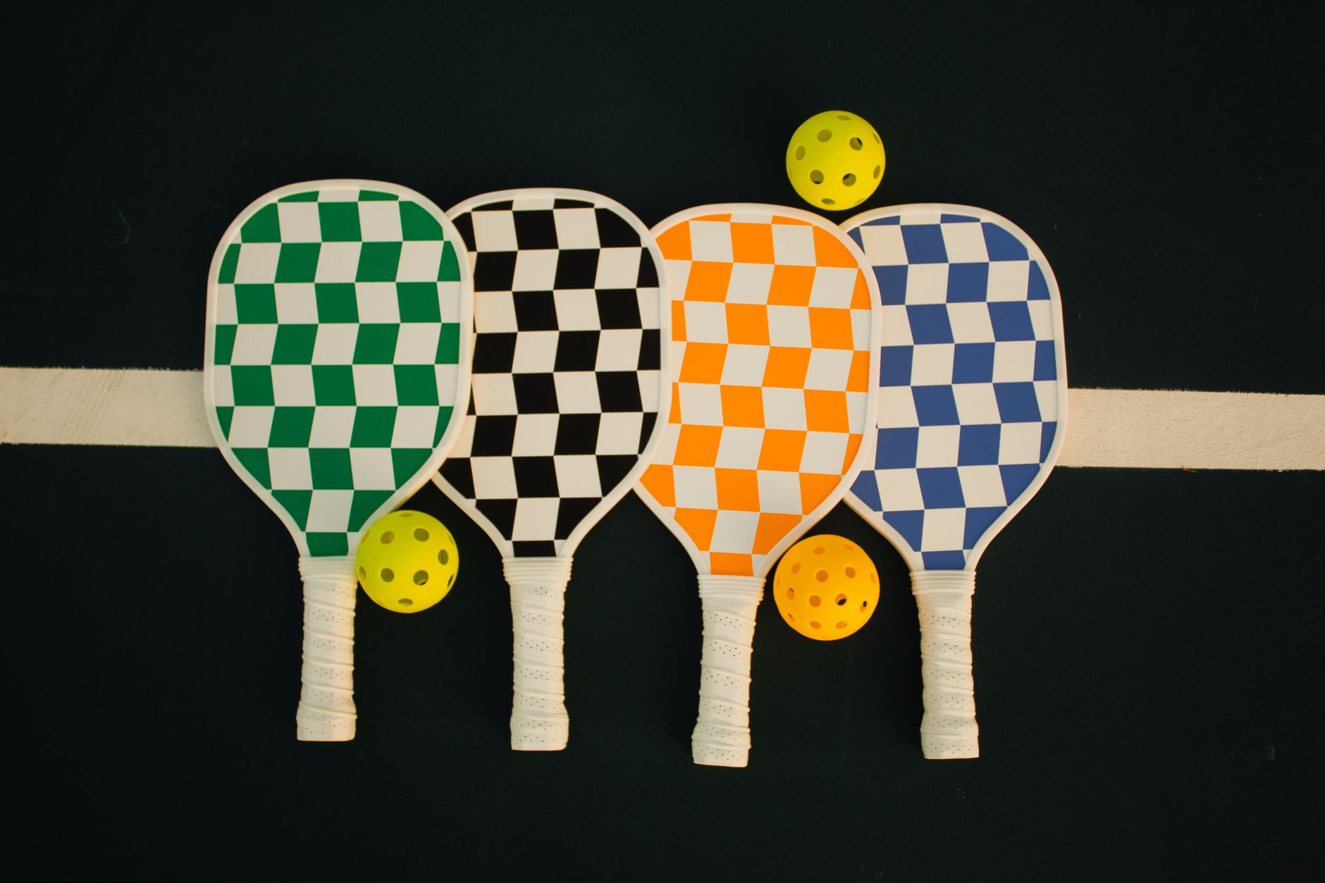 Four pickleball paddles in different colors arranged with a yellow ball on a court backdrop